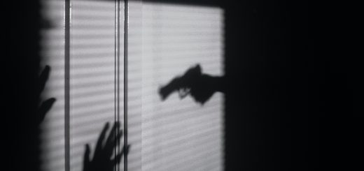 Two shadows. One is of a hand pointing a gun towards the second shadow. The second shadow looks like a person with their hands up.