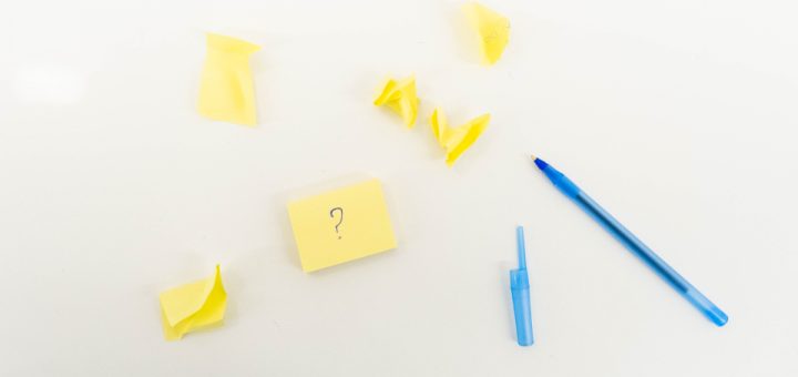 Several yellow post-it notes are scattered across a white desk. Some are crumpled up. One post-it note has a large blue question mark on it. There is an open blue pen in the bottom right of the image.